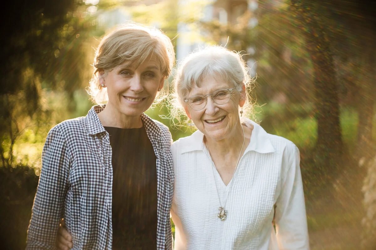 Two older women standing in a wooded area.