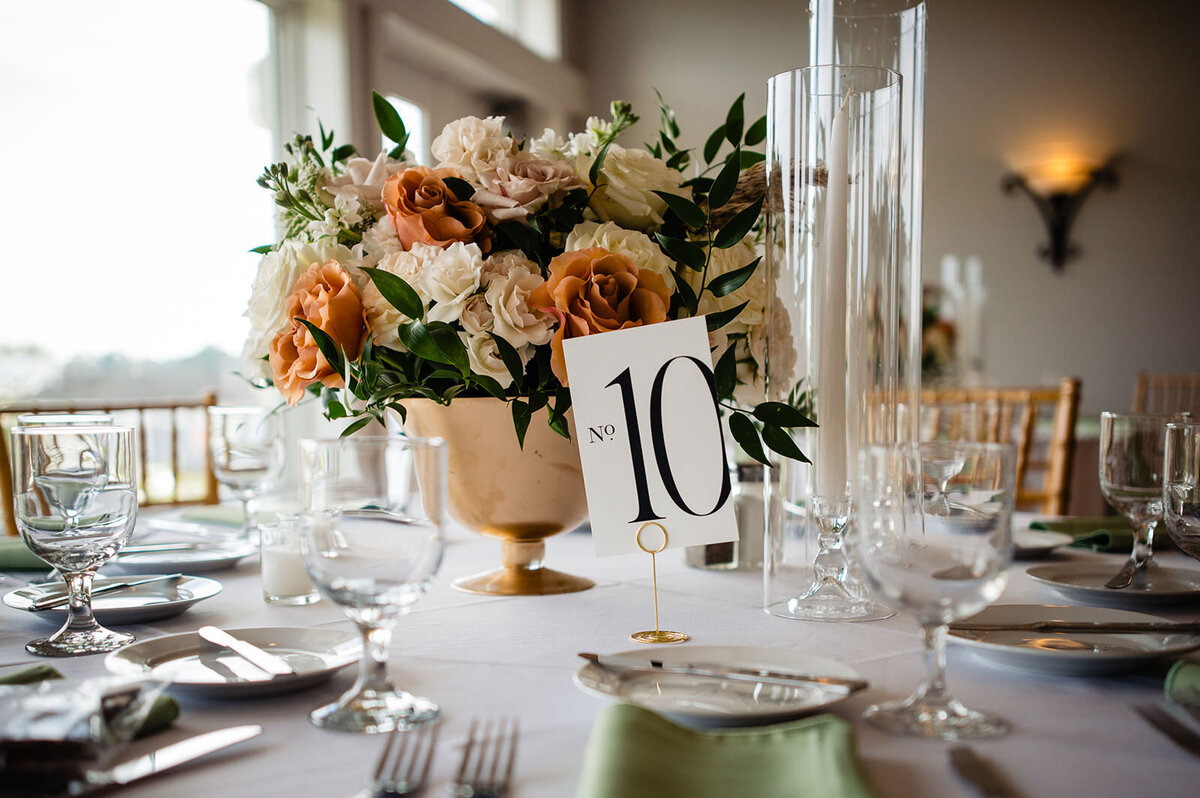 A wedding reception table elegantly set with glassware and a centerpiece of white and orange flowers with a table number sign.