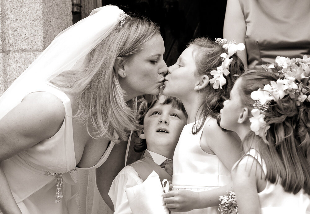 Bride with blonde hair leaning over to kiss a flower girl with flowers in her hair