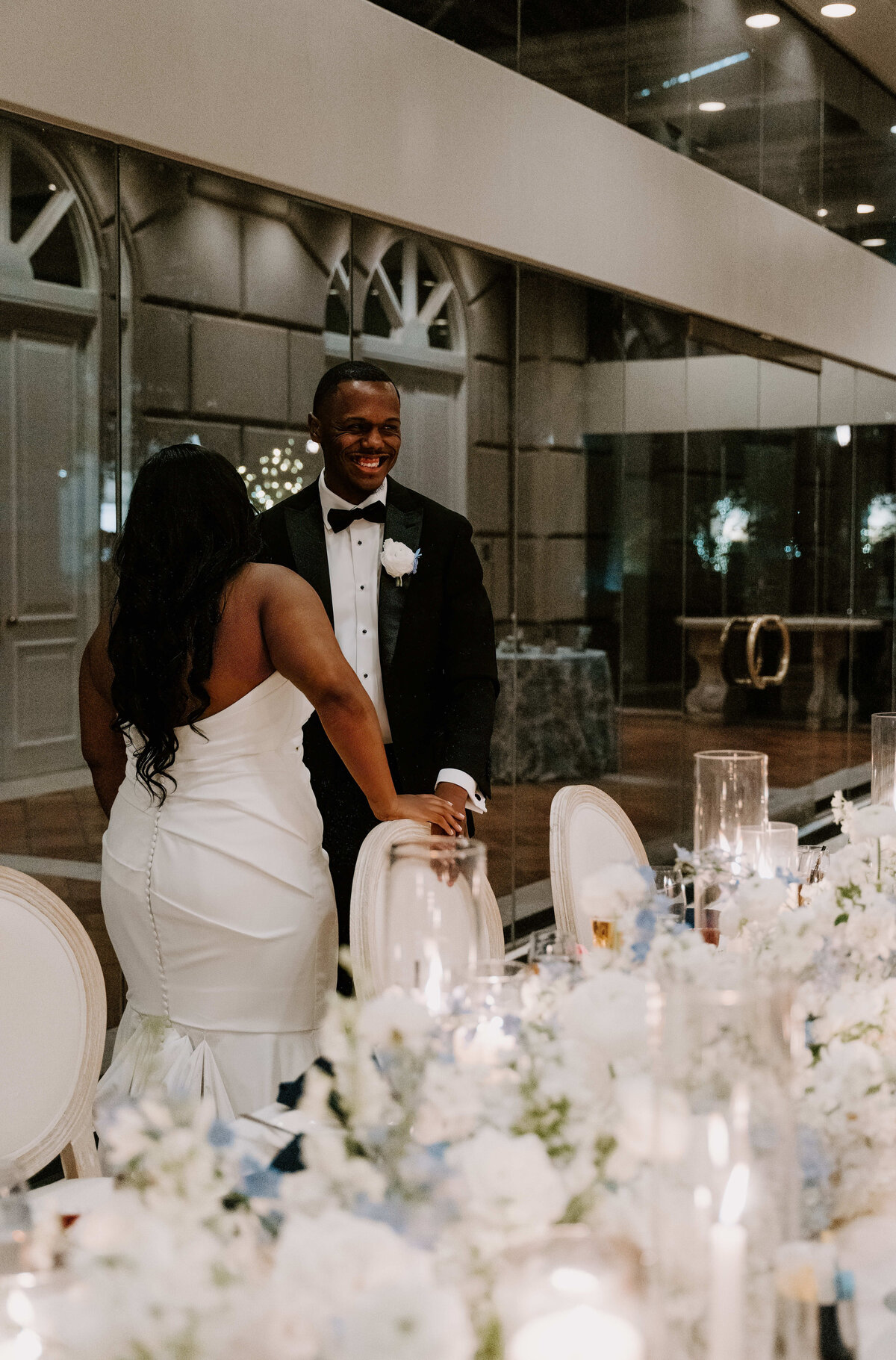 Bride in a white gown and groom in a tuxedo smile at each other at an elegant reception. The table is adorned with white and blue flowers and candles. Romantic atmosphere.