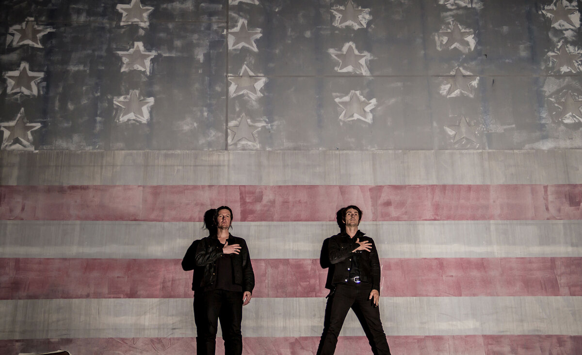 Musical duo portrait Walking Papers  standing hands over hearts giant American flag backdrop