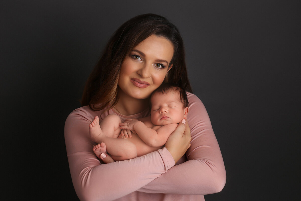 Mother wearing a pink top cradling her newborn near her face on a dark grey background.