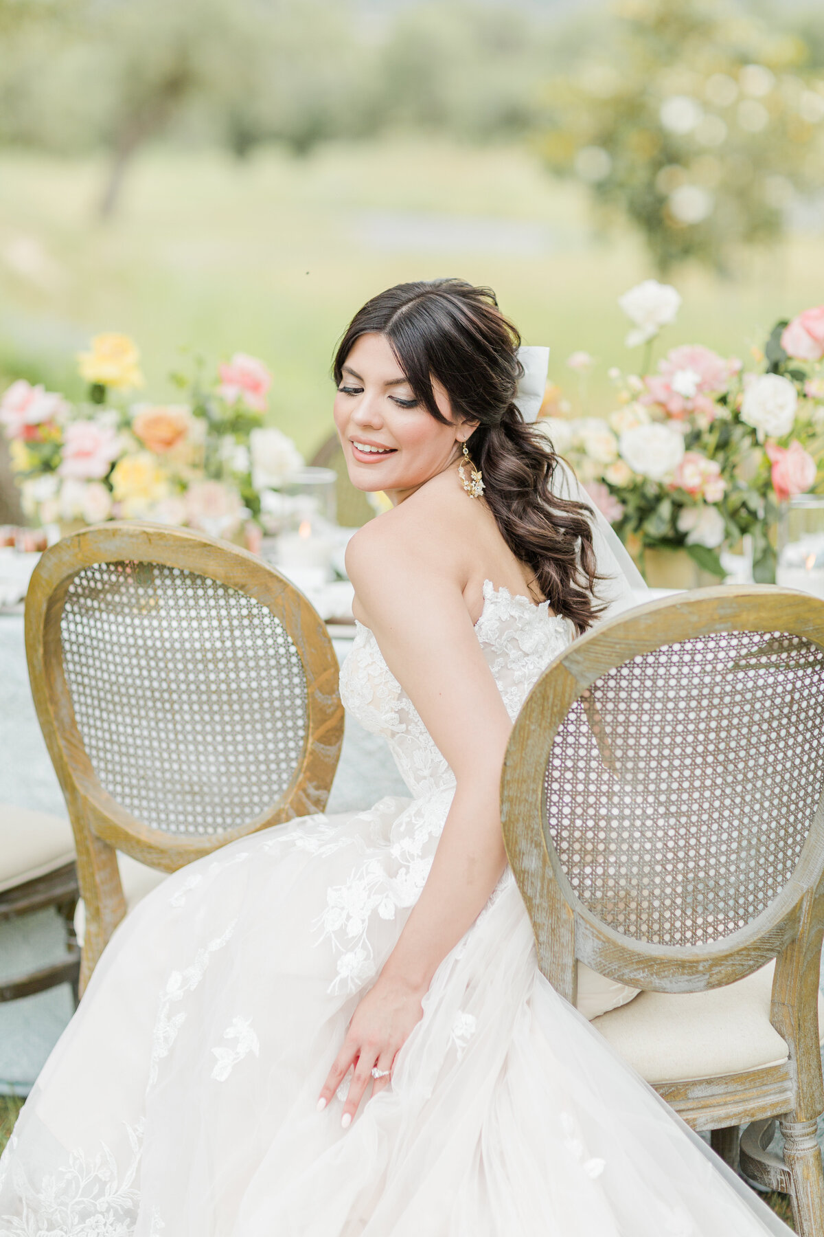 Bride is sitting on a chair for a casual portrait before her Boston wedding reception. the bride is sitting on the edge of the chair and looking over her shoulder smiling with her eyes closed for a joyful portrait. The table behind her is filled with pink, white, and yellow blooms. Captured by MA wedding photographer Lia Rose Weddings