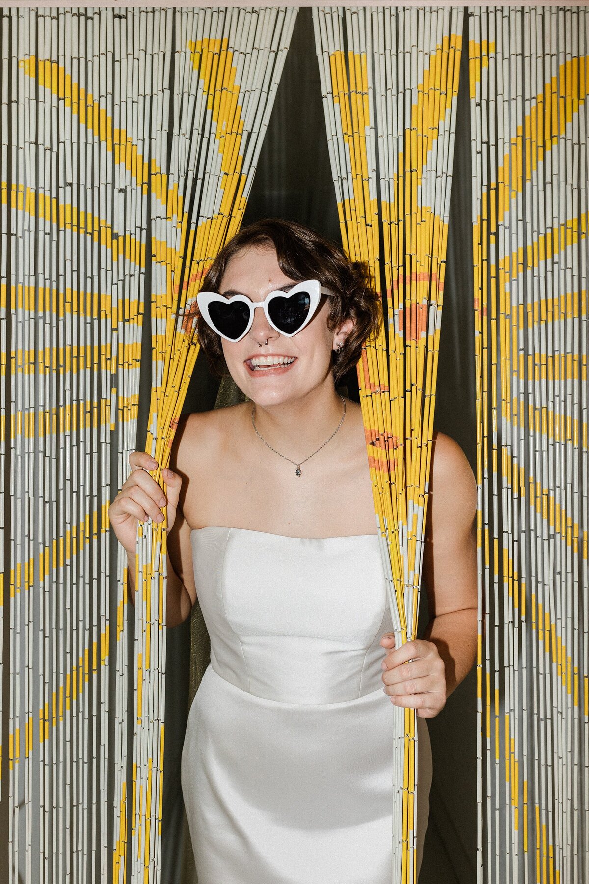 A portrait of a bride peering through a sun themed beaded doorway. The bride is wearing a simple yet elegant, sleeveless, white dress and white, heart-shaped sunglasses.