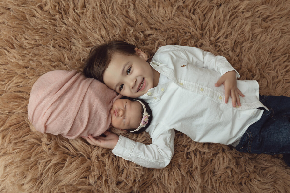 A toddler boy in a white shirt lays on a brown shag blanket head to head with his sleeping newborn baby sister during a NJ Newborn Photography session