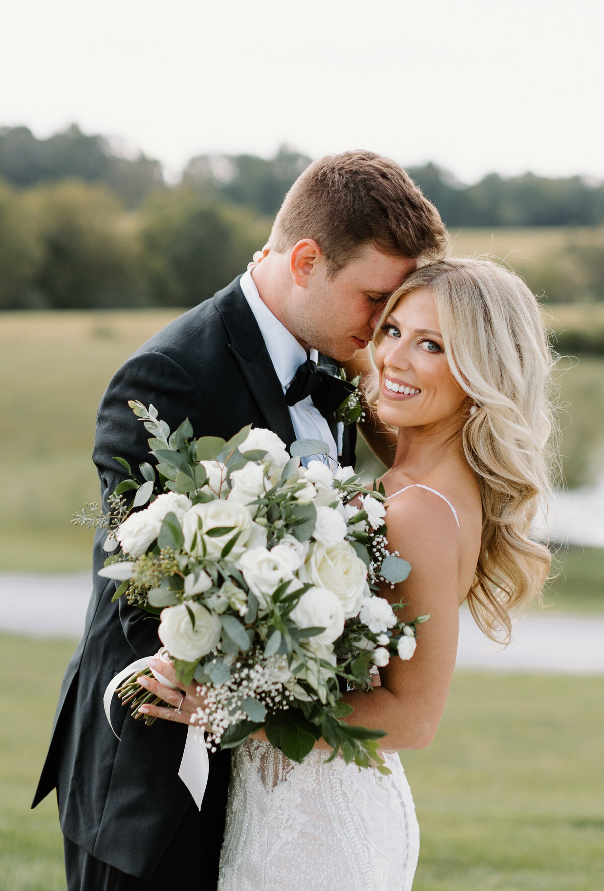 Bride smiling at camera as groom kisses her check, she's holding a large bouquet