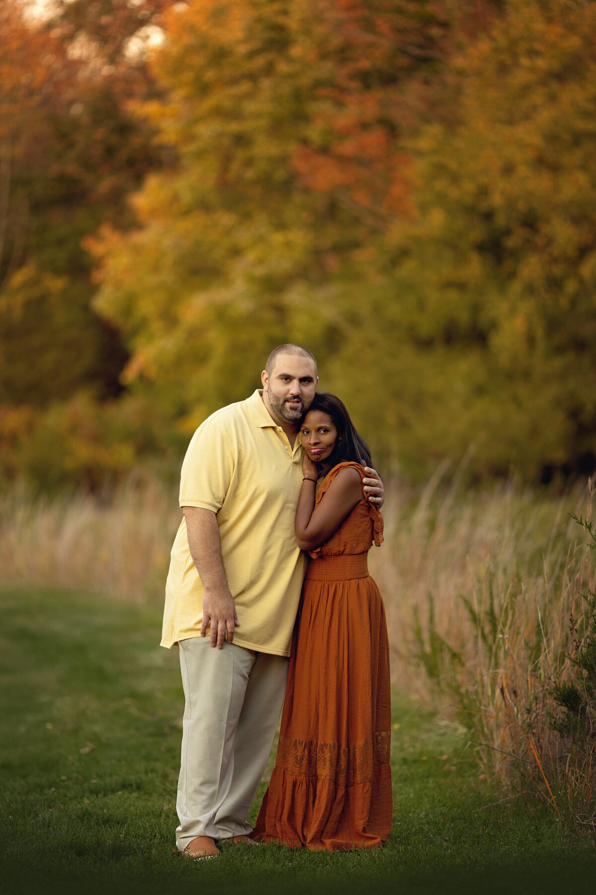 A mother in a red dress leans into the chest of her husband in a yellow shirt in a park at sunset