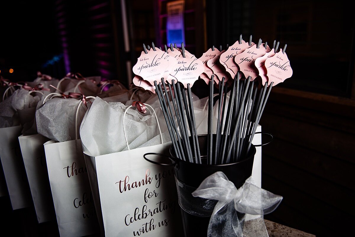 Wedding sparklers in a tall metal vase with paper thank you tags sit on a table next to gift bags for guests.