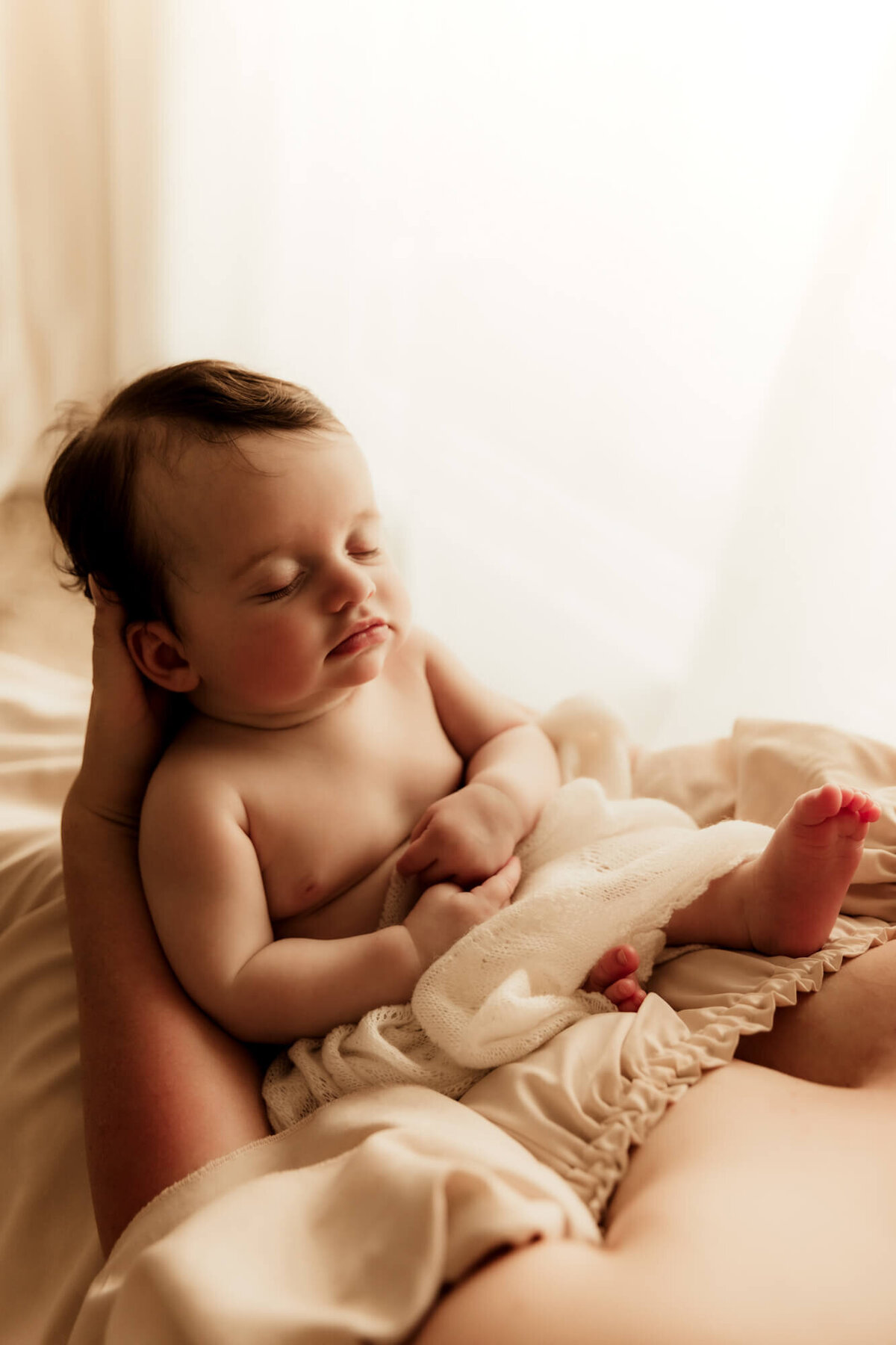 Mother holds her sleeping baby girl in her lap while wearing a cream dress for a studio photo in OKC.