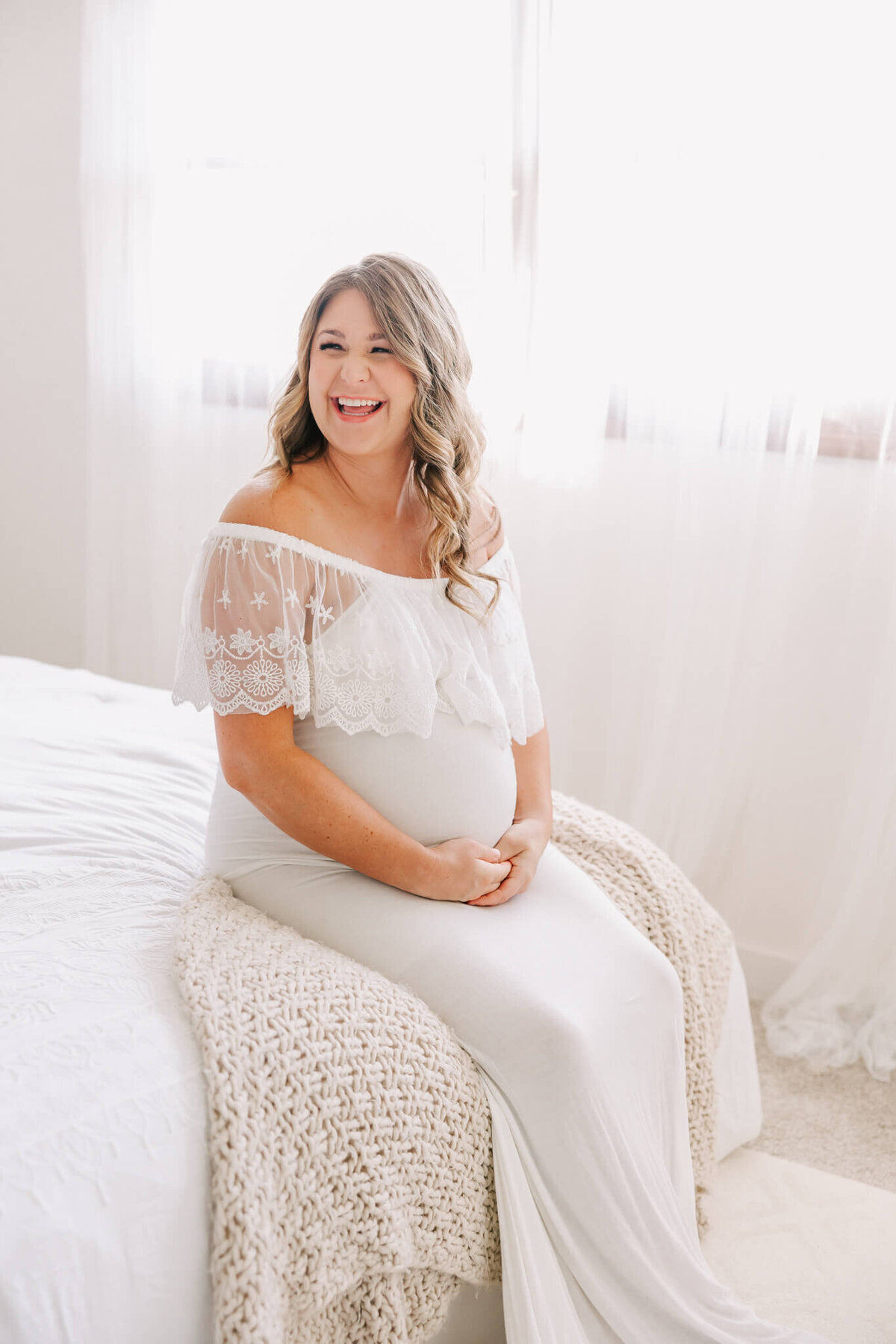 beautiful portrait of a woman smiling and laughing while sitting on a bed for her maternity session