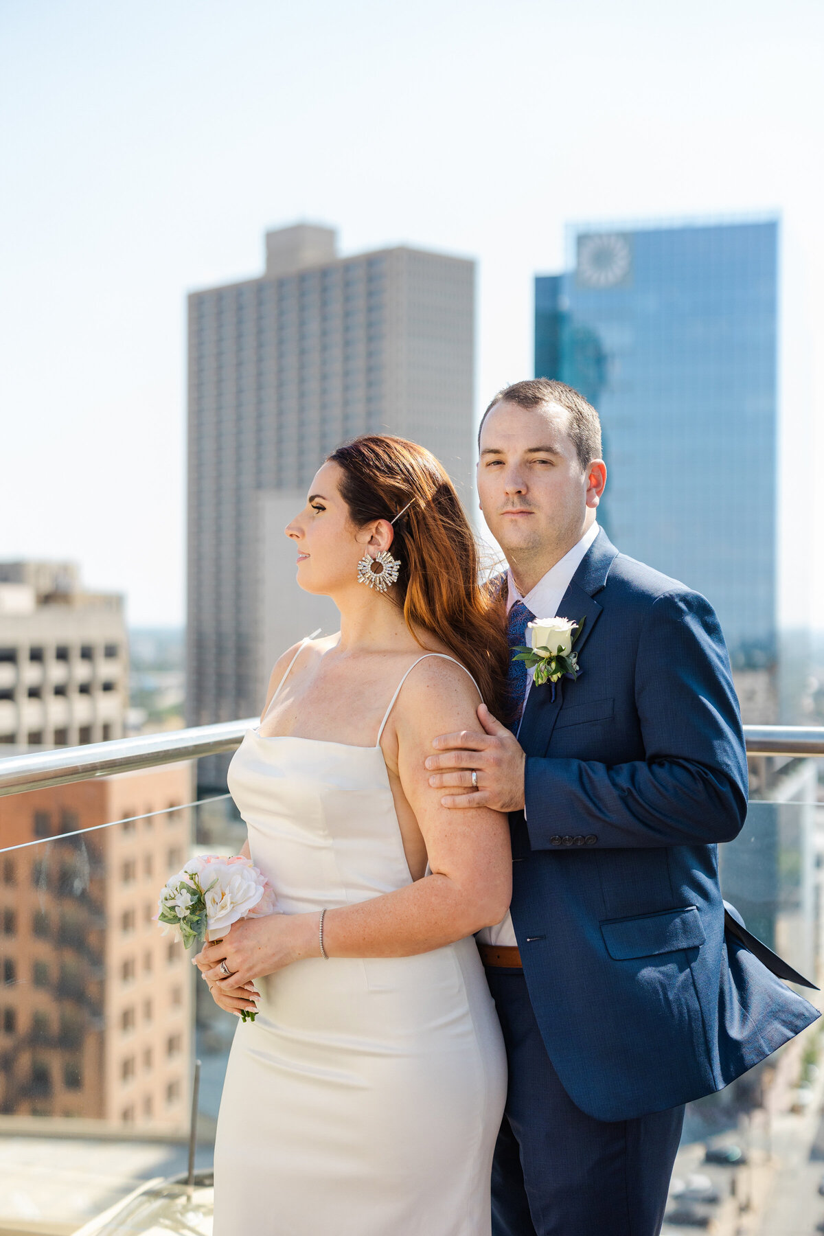 Portrait of a couple posing on the roof of a building in downtown Fort Worth after their elopement ceremony at the Tarrant County Courthouse in Fort Worth, Texas. The bride is on the right and is wearing a sleeveless, white dress and is holding a bouquet. The grooms stands behind her on the right and is wearing a navy suit with a boutonniere. The Fort Worth skyline can be seen in the background.
