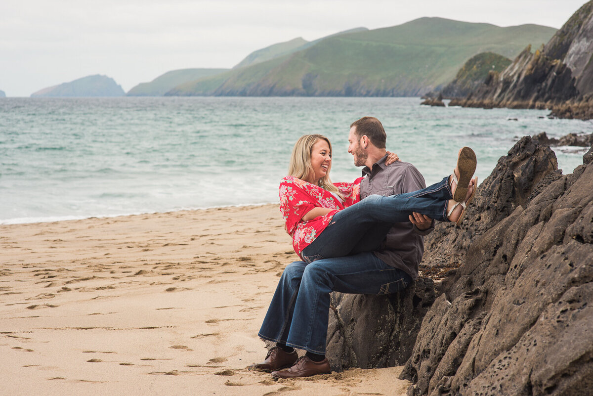 engaged couple wearing denim sitting on a rock on the beach in Dingle having fun