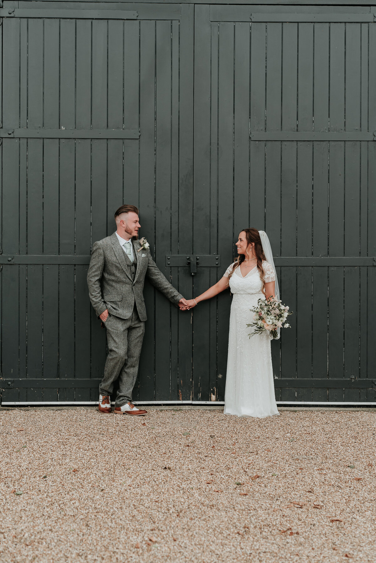 Bride in beautiful lace dress and Groom in tweed grey suit hold hands leaning on black wooden barn doors at Bury Court Barn