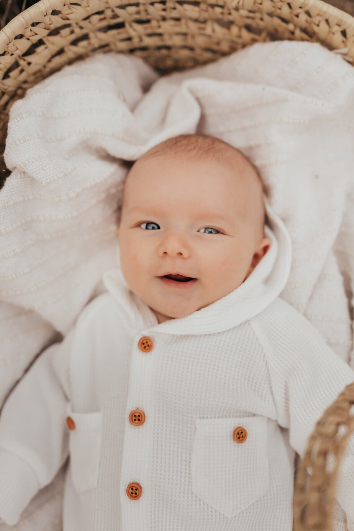 little baby boy smiles while wearing a white romper and laying in moses basket