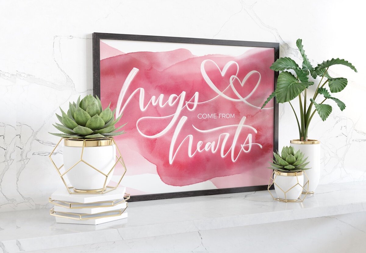 Framed pink watercolor painting with hearts and hand lettered words "hugs come from hearts"
