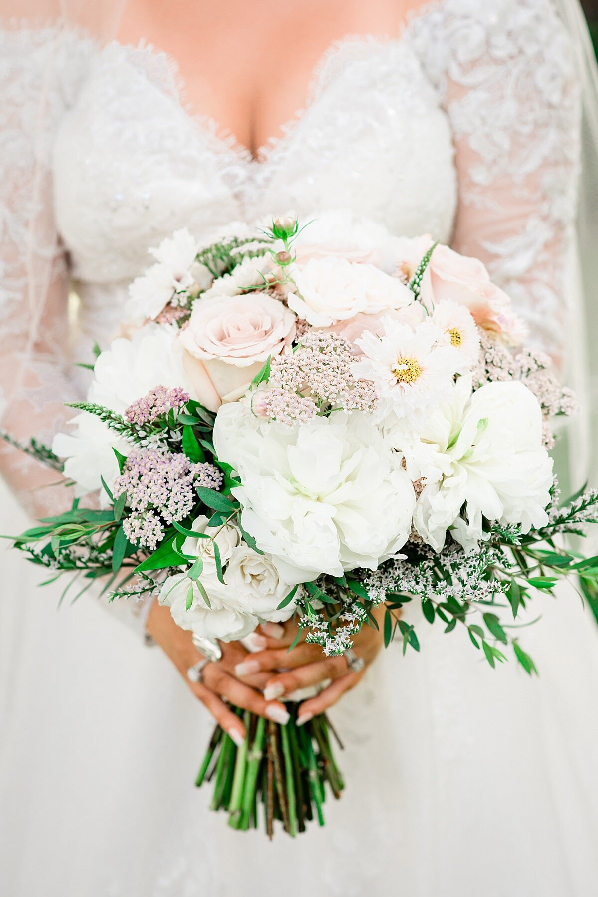 Detail of the bridal bouquet. The bride is wearing a long sleeved lace wedding dress with a deep v neckline. The bridal bouquet is all white with hints of greenery, light pink rice flower, white peonies, white roses,  blush roses, and other white flowers.