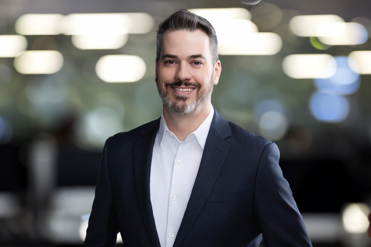 Professional headshot of a white male with salt-and-pepper hair and beard, wearing a navy suit and white dress shirt. He has a confident, friendly expression with a slight smile and direct eye contact. Photographed against a modern, softly blurred office background with bokeh lighting for a polished, executive look. Captured at Janel Lee Photography in downtown Cincinnati, Ohio—specializing in high-end headshots for speakers, executives, and business professionals.