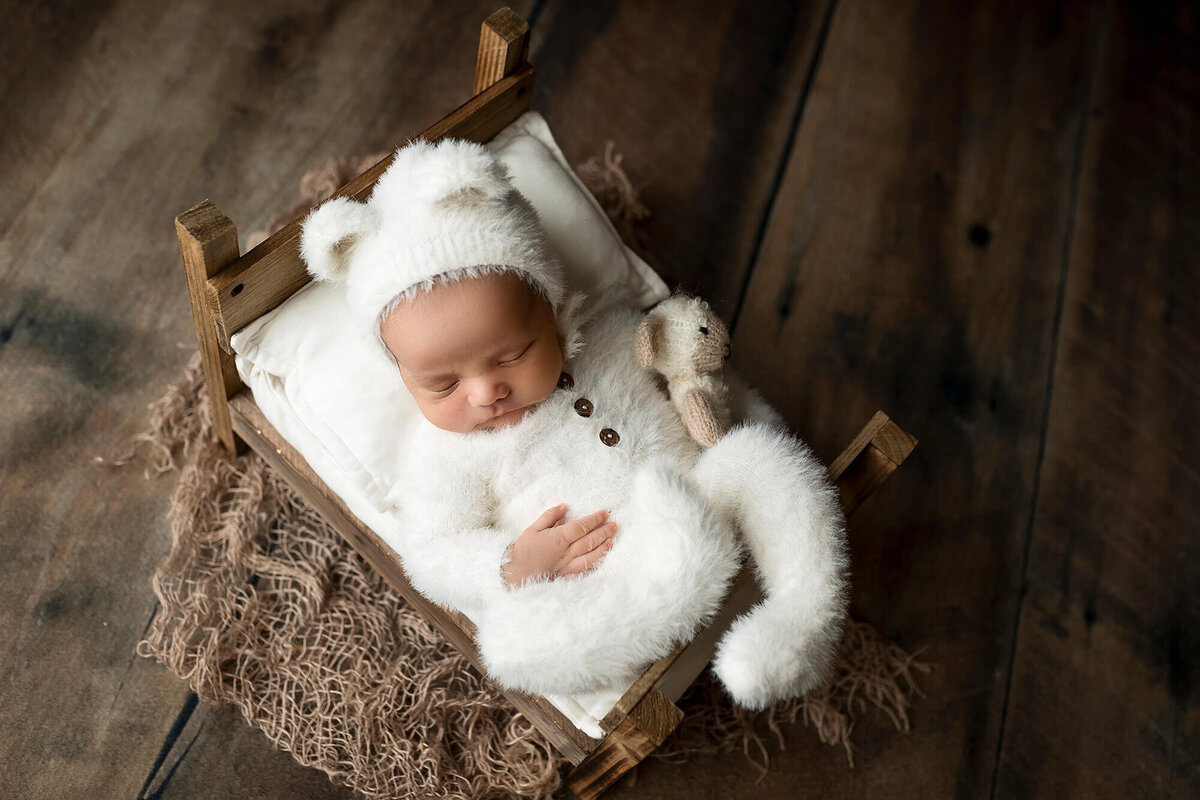Newborn boy with white bear suit sleeping on a tiny bed.