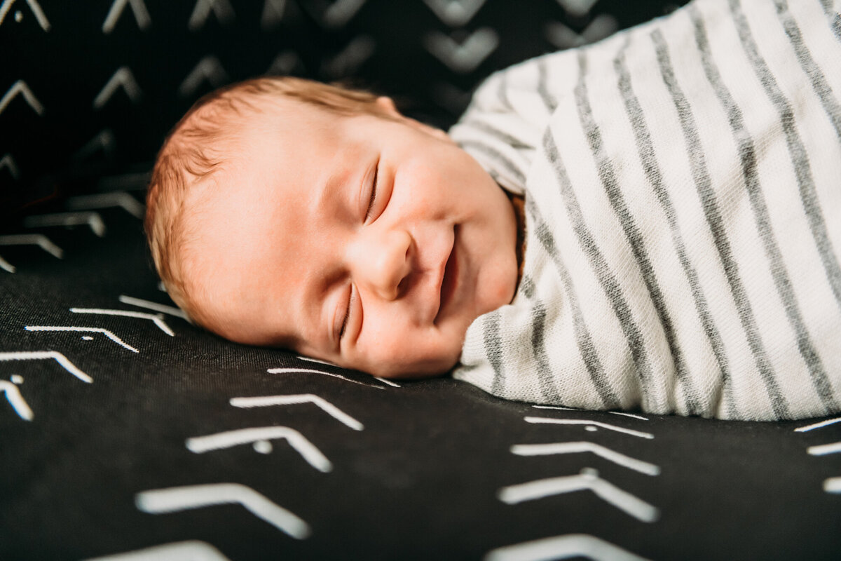 Newborn Photographer, Baby smiling and sleeping on a black cushion while he's swaddled.