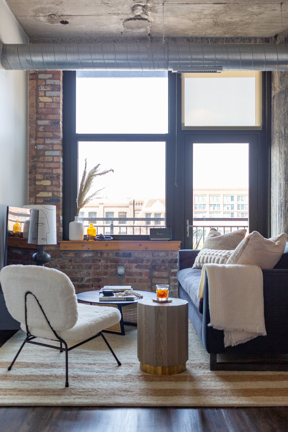 Industrial loft featuring brick walls and concrete columns with blue velvet sofa, arched cane divider screen, striped jute rug, and furry white lounge chair