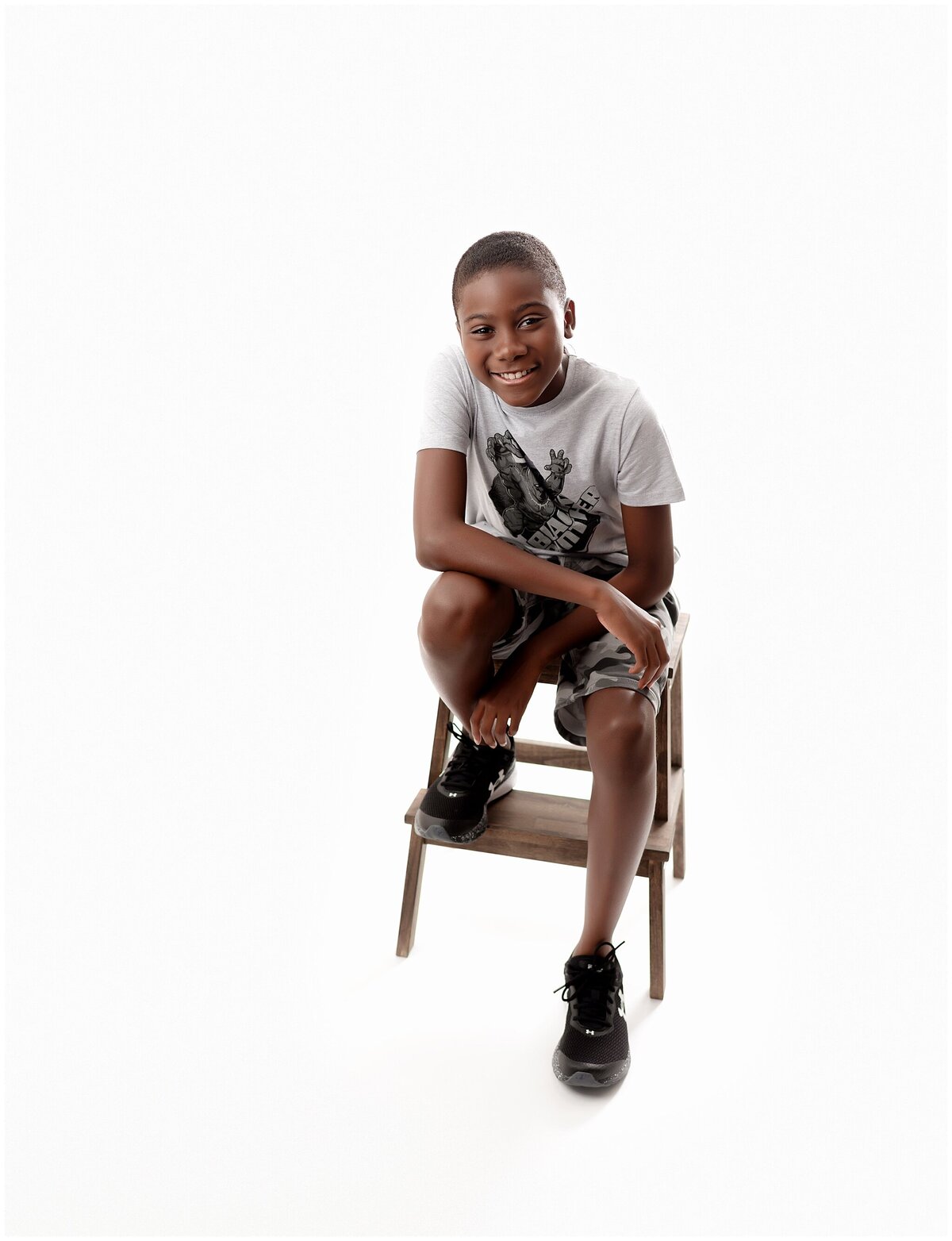 A portrait of a boy sitting on a stool with one leg on one step and the other foot on the floor. He is leaning one arm on his leg and looking at the camera.