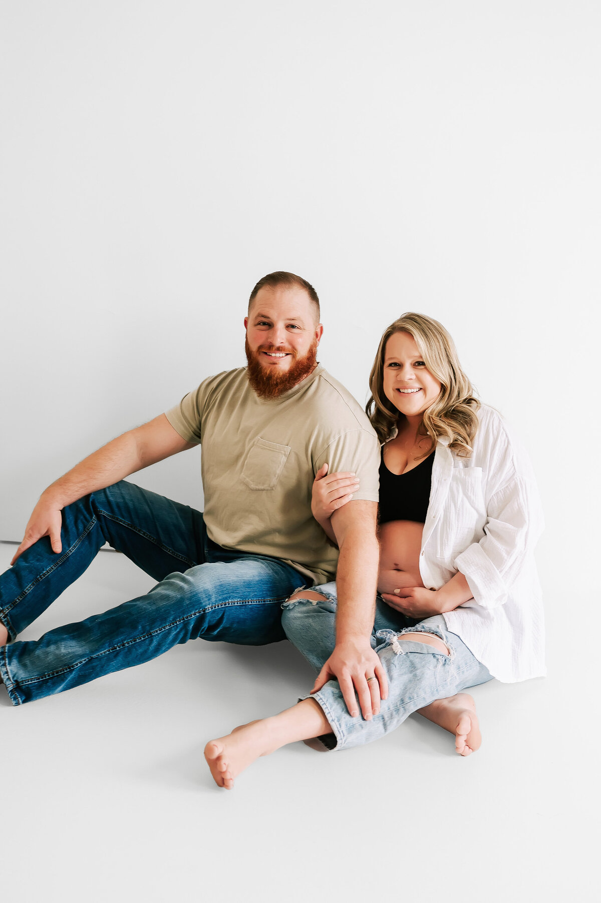 husband and wife sitting on ground smiling holding pregnant belly enjoying maternity pictures in Springfield MO