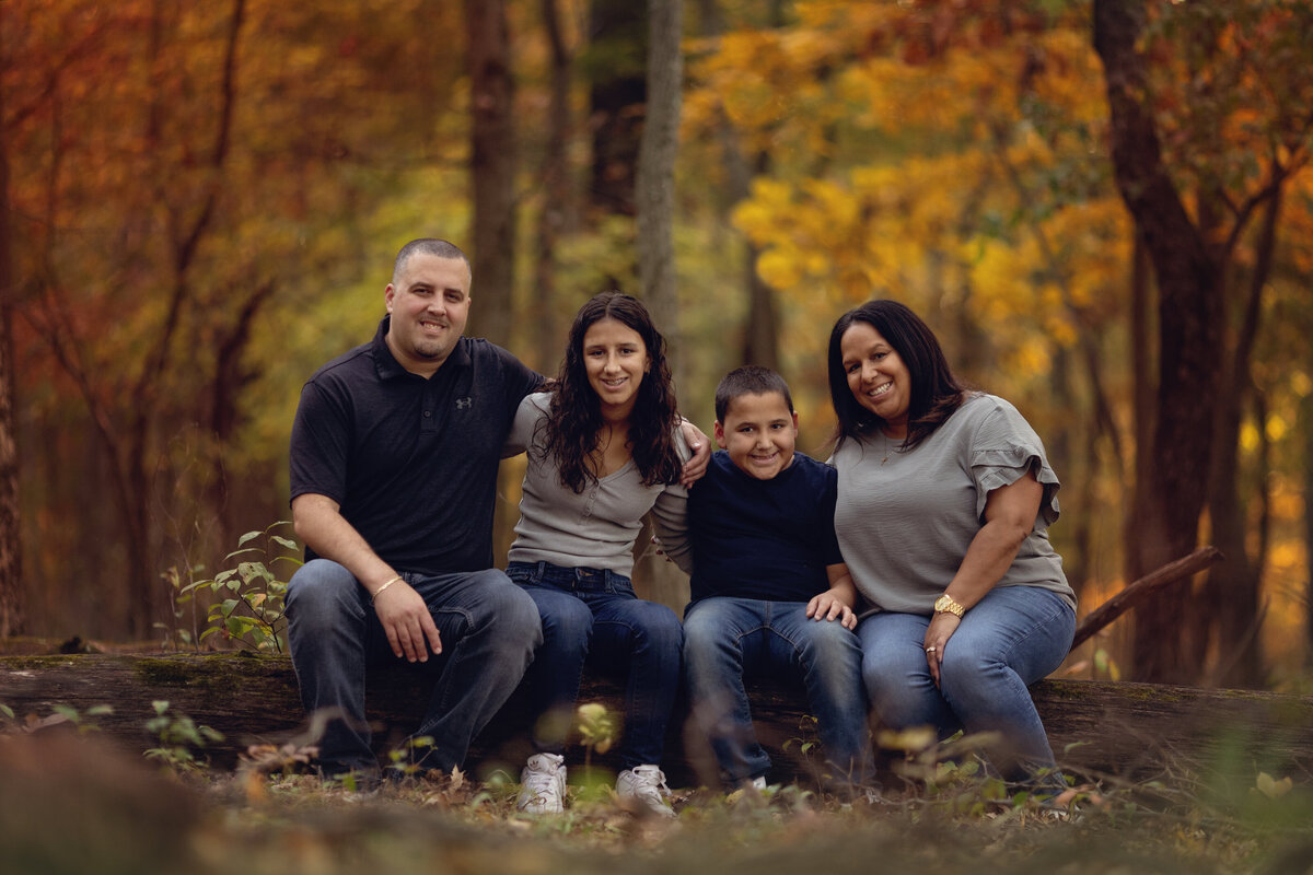A mother and father sit on a fallen log with their two teen children with arms around each other during a session with a New Jersey Family Photographer