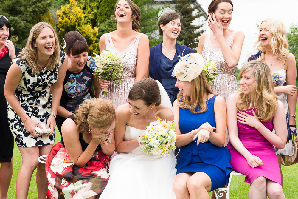 Bride in A-line wedding dress holding white flower bouquet sitting with friends in garden laughing