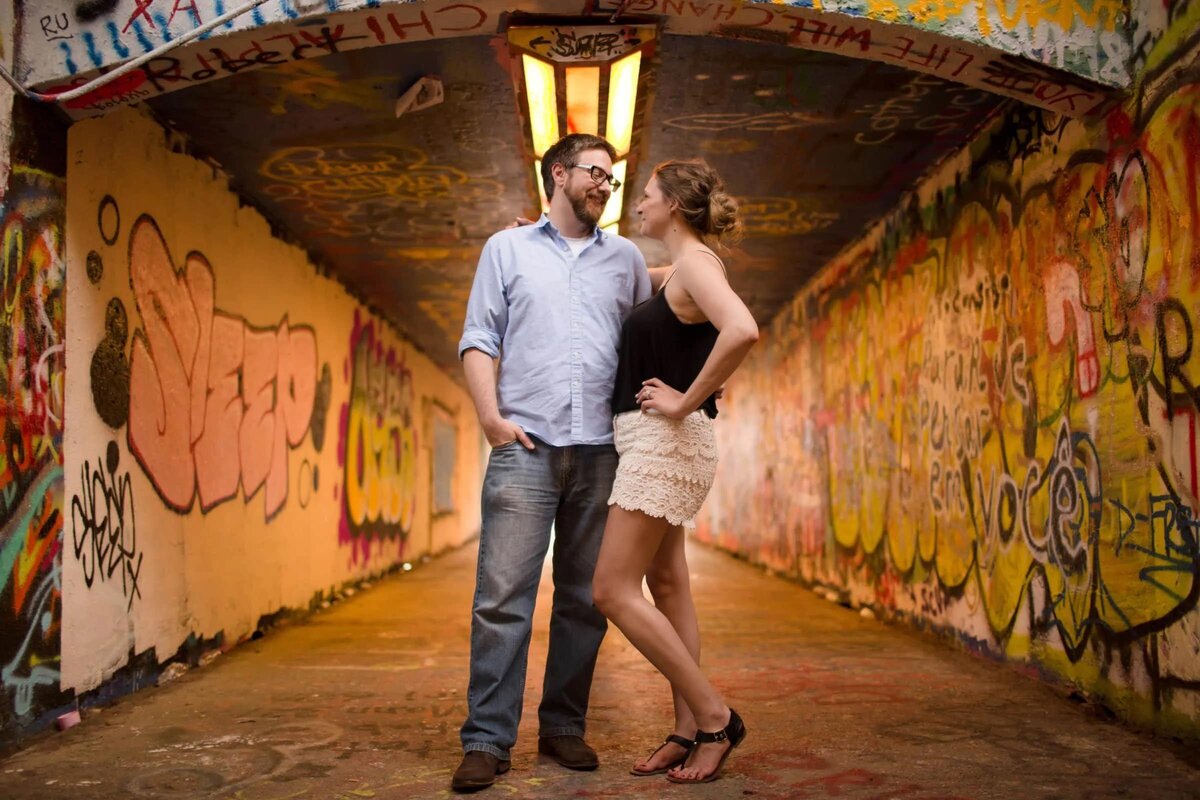 A couple stands in a graffiti-covered tunnel, sharing a laugh, with warm light emanating from behind