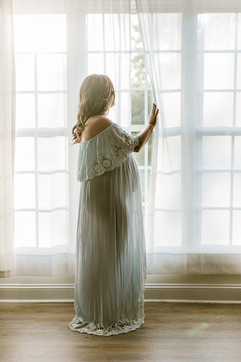 pregnant mom stands by window and light shines through her dress silhouetting her round stomach