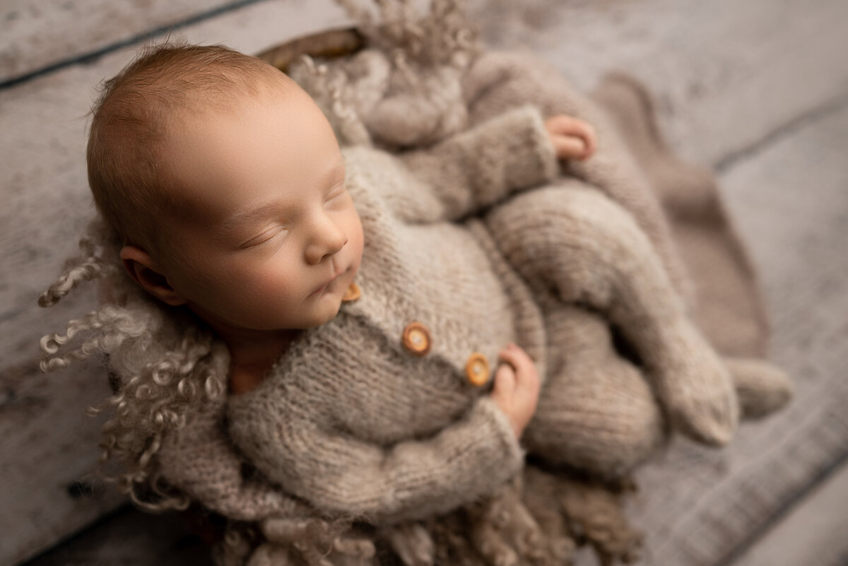 Katie Marshall, New Jersey's best newborn photographer captures a fine art newborn photo. Baby is in a knit taupe one-piece outfit  sleeping in a bowl. Baby's legs are folded and his hands resting on his belly.