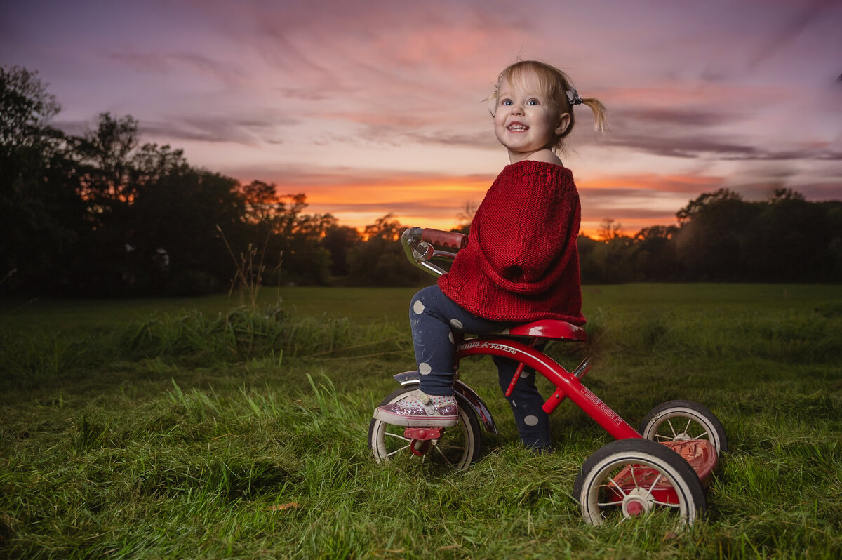 Young girl in red on tricycle with colorful sunset behind near Boston