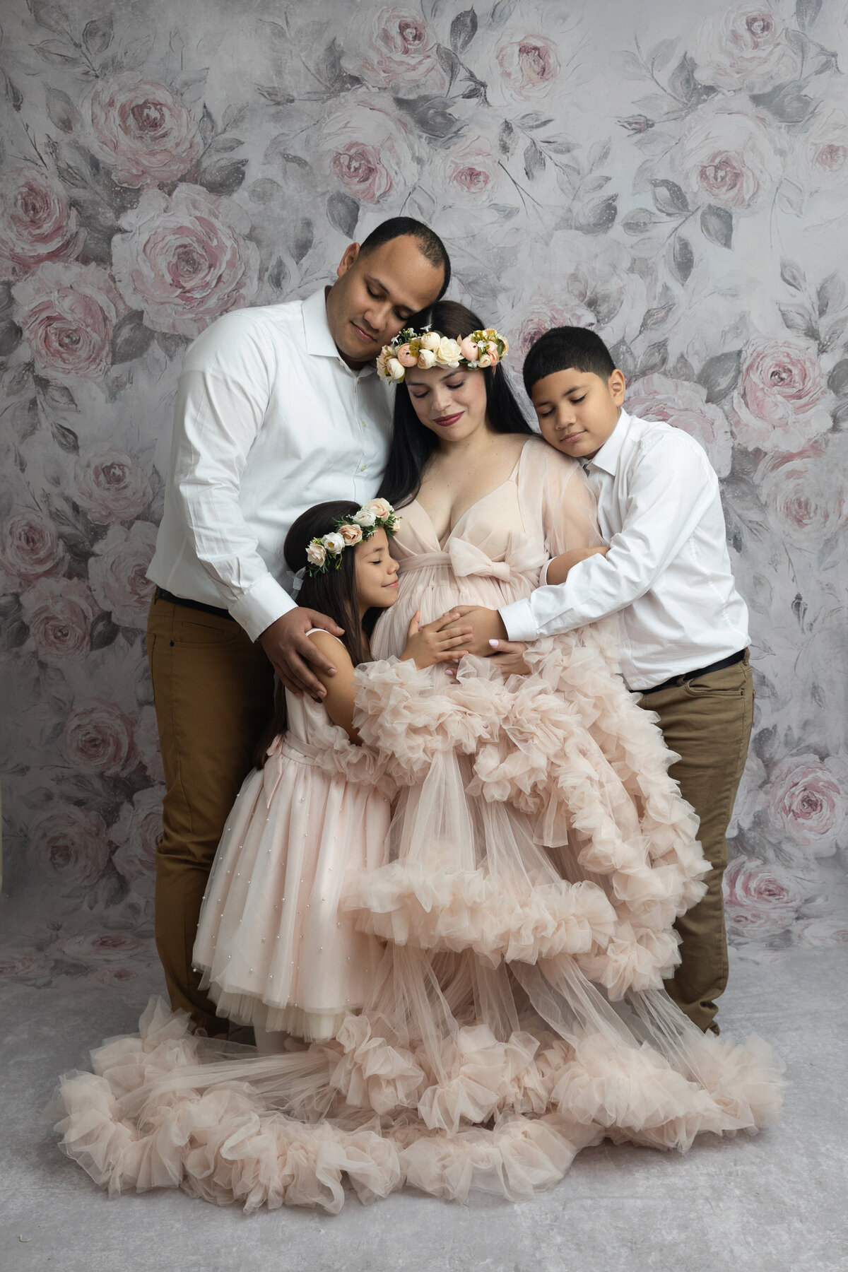 A father and his young son and daughter hug their pregnant mother in a studio with a floral print background as posed by a New Jersey Maternity Photographer