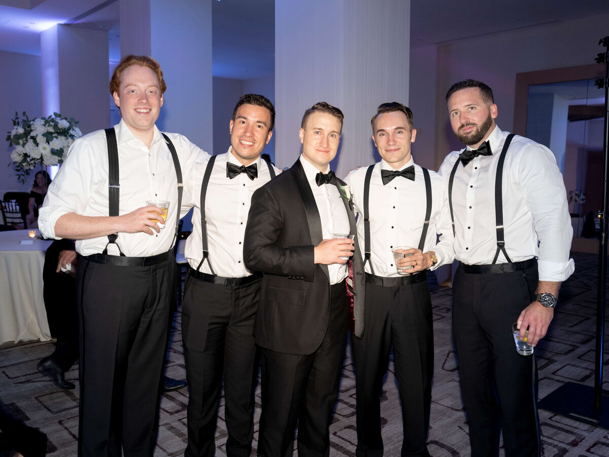 Five men wearing formal attire, including bow ties and suspenders, pose together at an indoor event. They hold drinks and smile at the camera. The room is softly lit, with floral arrangements visible in the background.