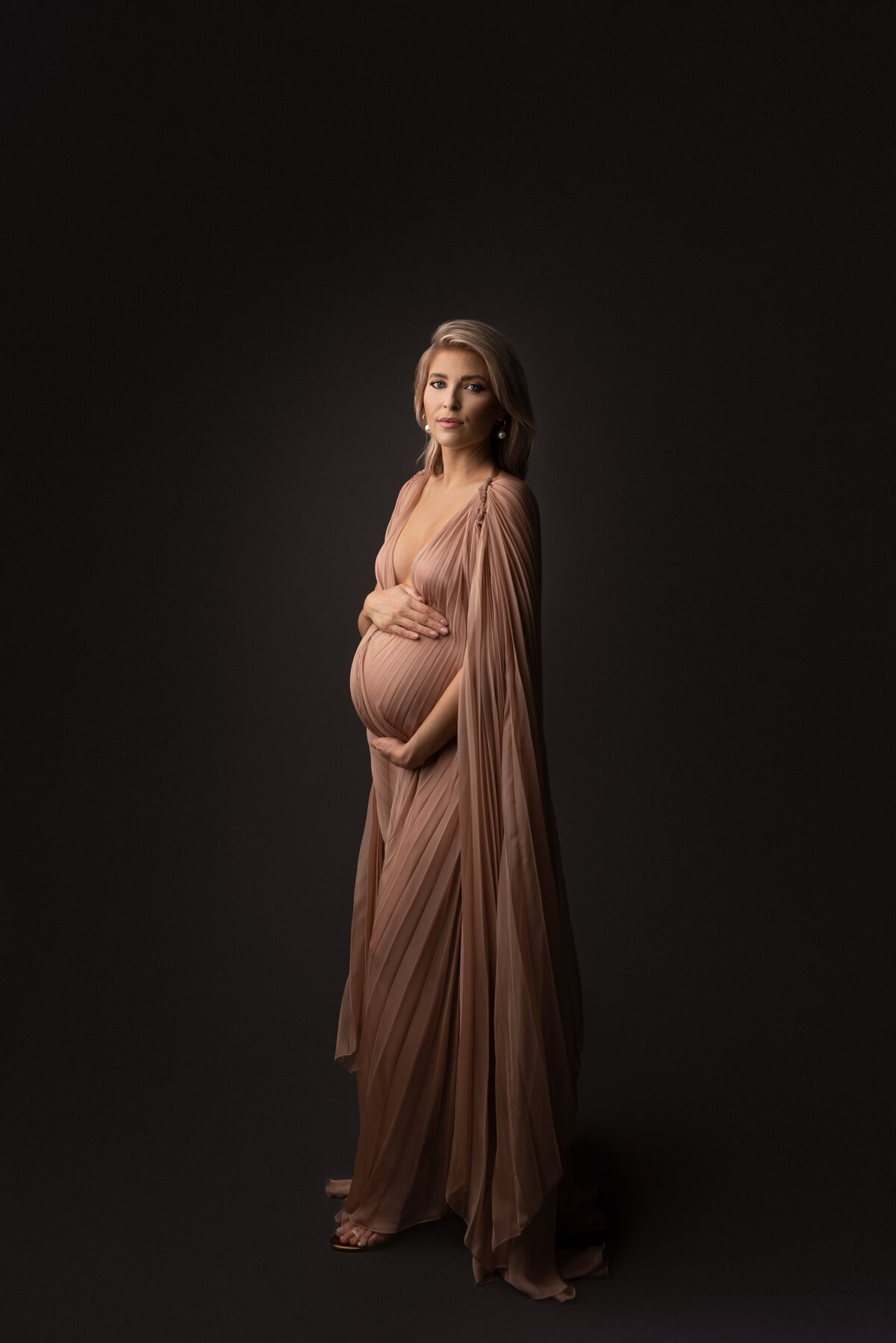 Woman poses for fine art maternity photos with New Jersey's best maternity photographer Katie Marshall. Woman in a floor-length dusty rose maternity gown with floor-length caped sleeves stands angled to the camera. One hand is above her baby bump, the other underneath. She is looking over her shoulder at the camera. Light envelops her face; her back is shadowed creating depth.