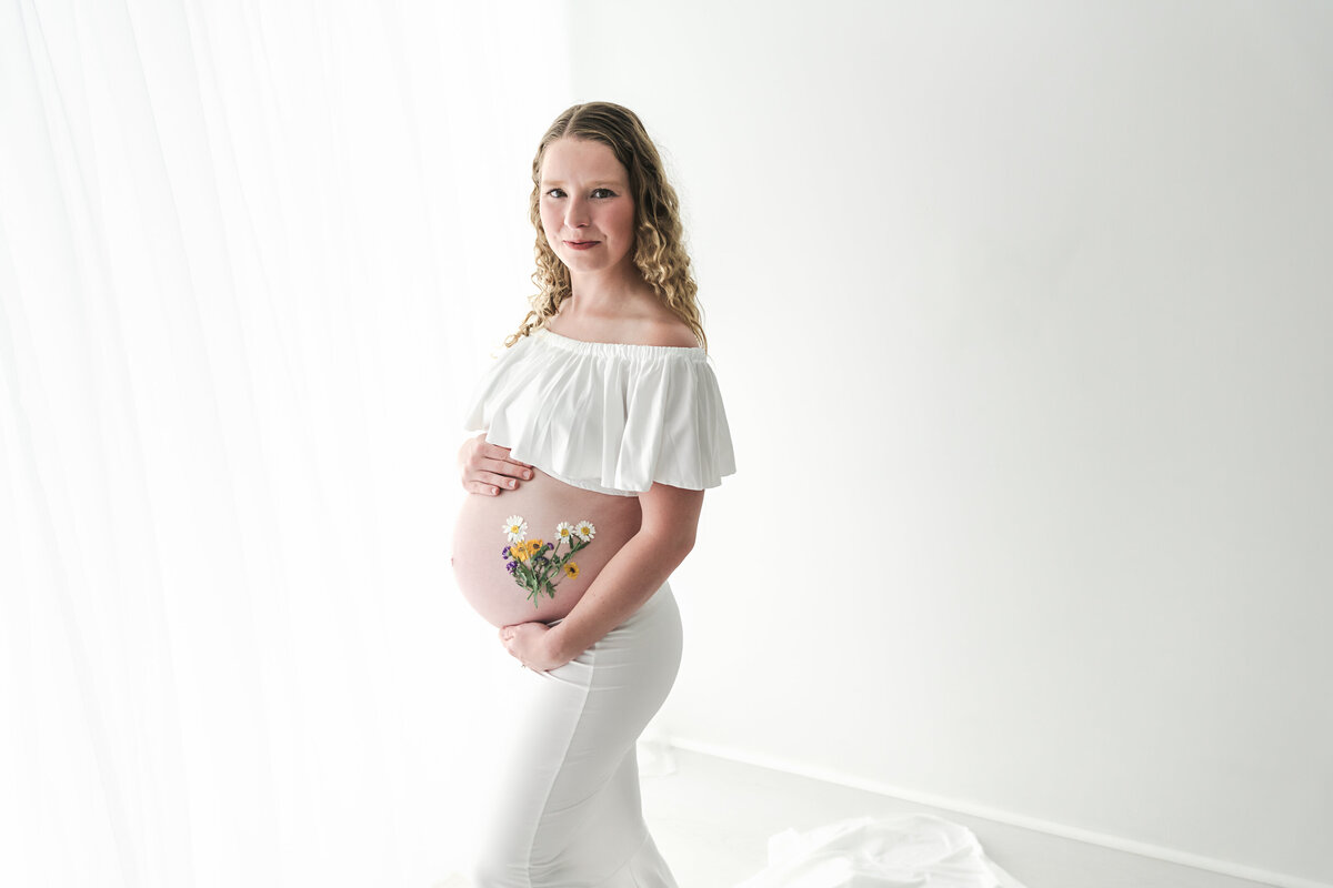 Expectant mother in a white outfit cradling her belly with a gentle smile, radiating happiness and anticipation.  She has flowers on her belly.  Taken by Fig and Olive Photography, a Minneapolis Maternity Photographer.