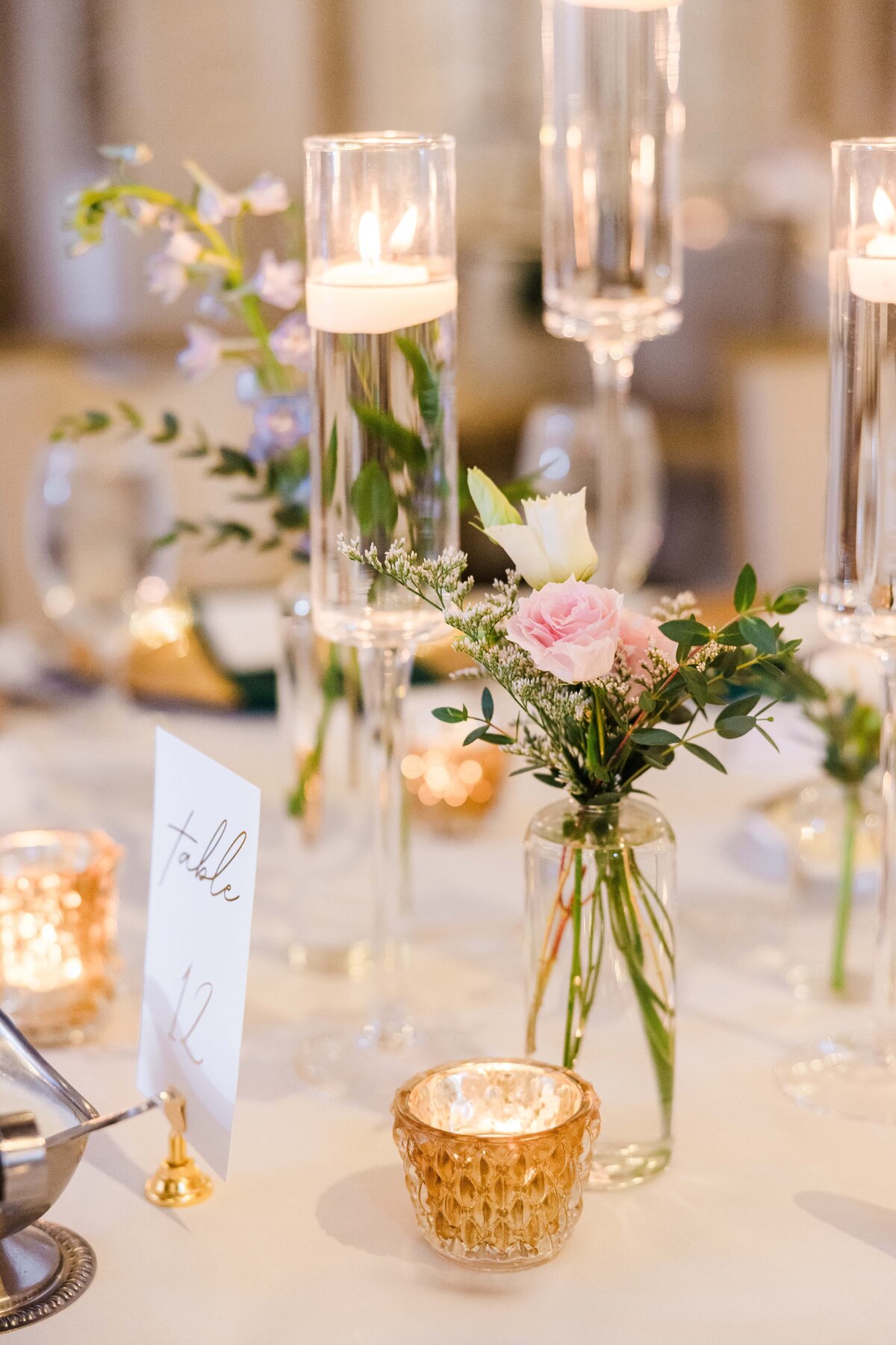 Elegant wedding table setting featuring candles in tall glass holders, a small bouquet with pink roses, and a table number card, meticulously arranged by a renowned wedding planner from Des Moines.