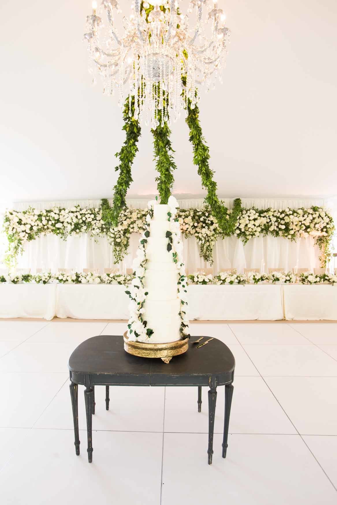 White wedding cake on table beneath a crystal chandelier and green garlands