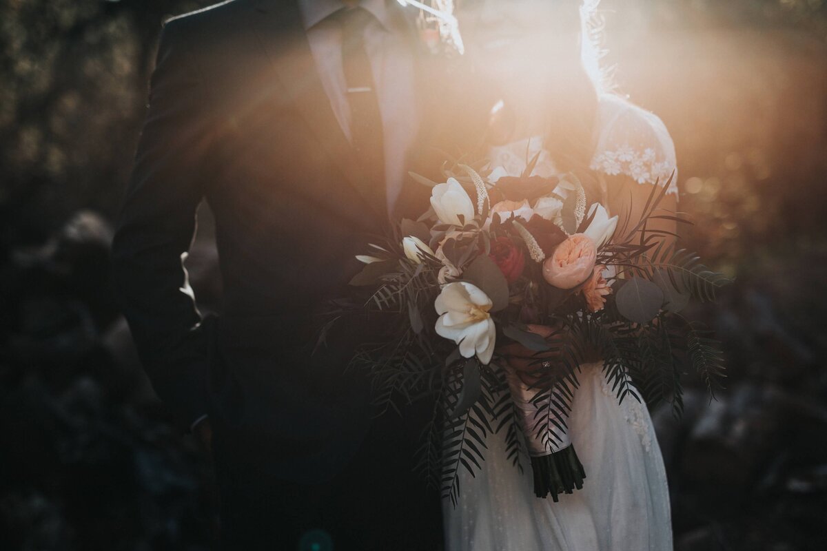 A couple on their wedding day. The groom in a black suit and the bride wearing a white lacy dress holding a green, white and red bouquet