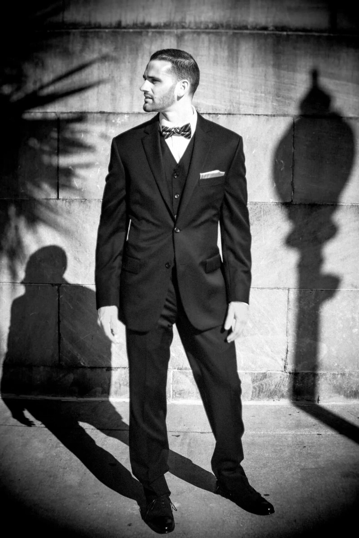 Black and white photo of a groom in formal attire, standing confidently with striking shadows casting patterns behind him