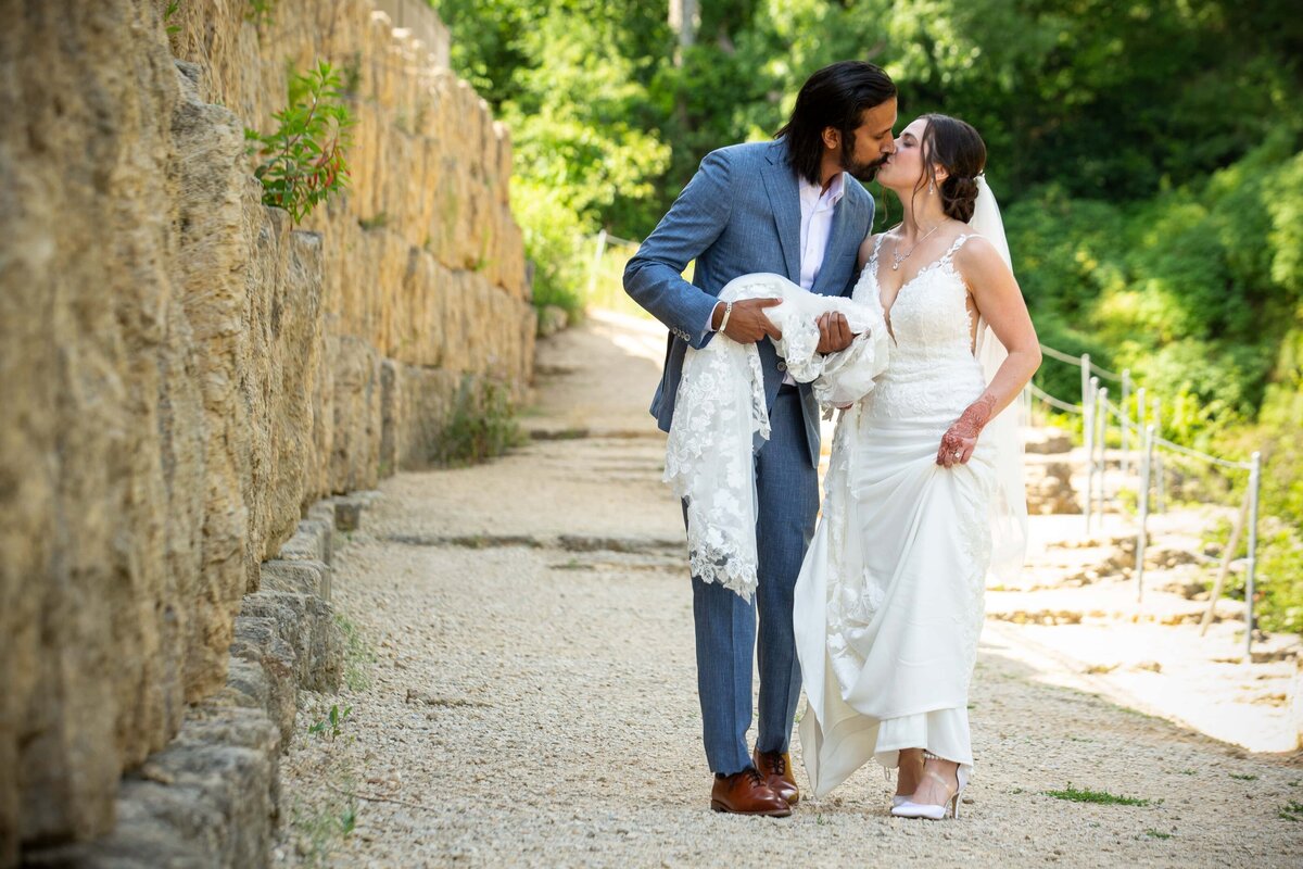 A couple in wedding attire sharing a romantic moment on a stone-paved path beside a rock wall, with the groom holding the bride's dress during their Iowa wedding.