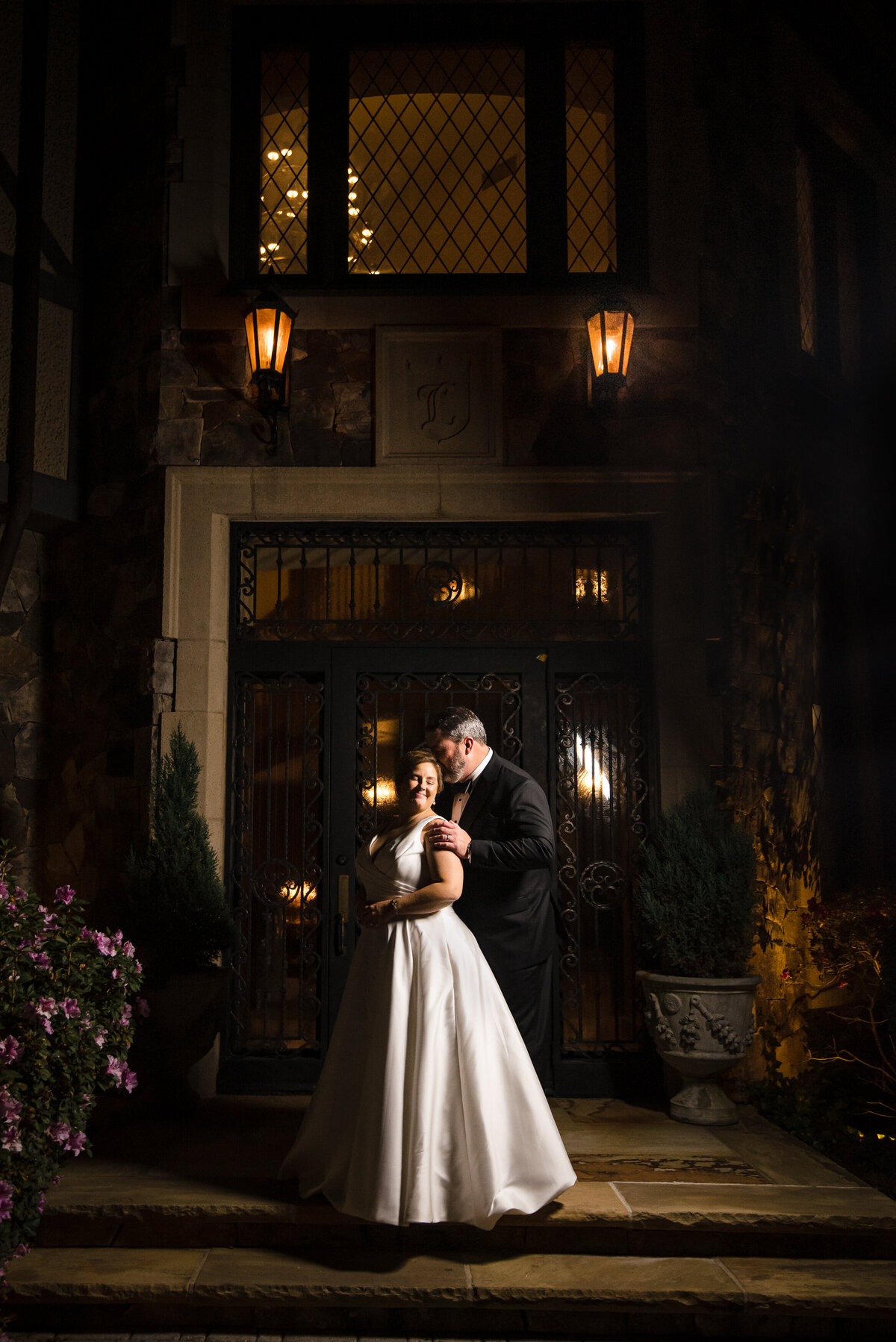 Nighttime-portrait-of-groom-kissing-his-bride's-temple-in-front-of-the-doors-of-The-Club-at-Longview-by-Charlotte-wedding-photographers-DeLong-Photography