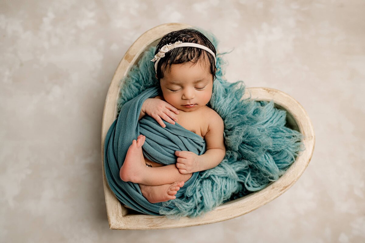 newborn girl is wrapped in a teal wrap and posed in a cream heart bowl in a charlotte studio