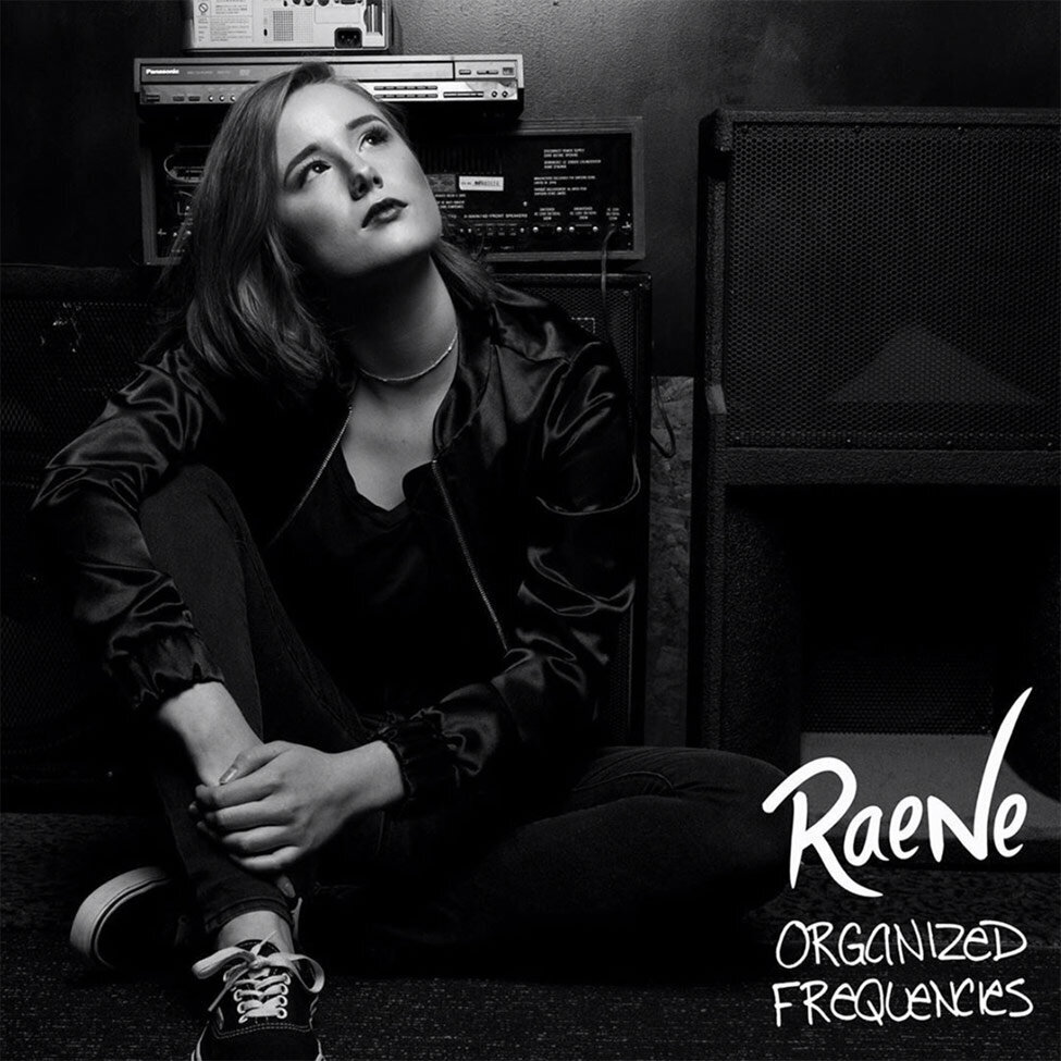 Single Cover Title Organized Frequencies Artist Raene black and white portrait sitting on floor with arms around bent knee hands clasped together while looking up amplifiers and speakers behind her