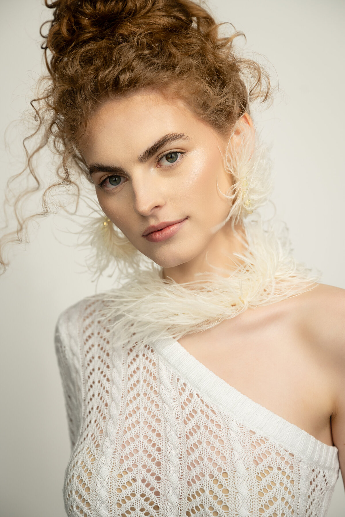 A portrait of a young woman wearing her hair up with loose curls looking straight to the camera. She is wearing a feather necklace