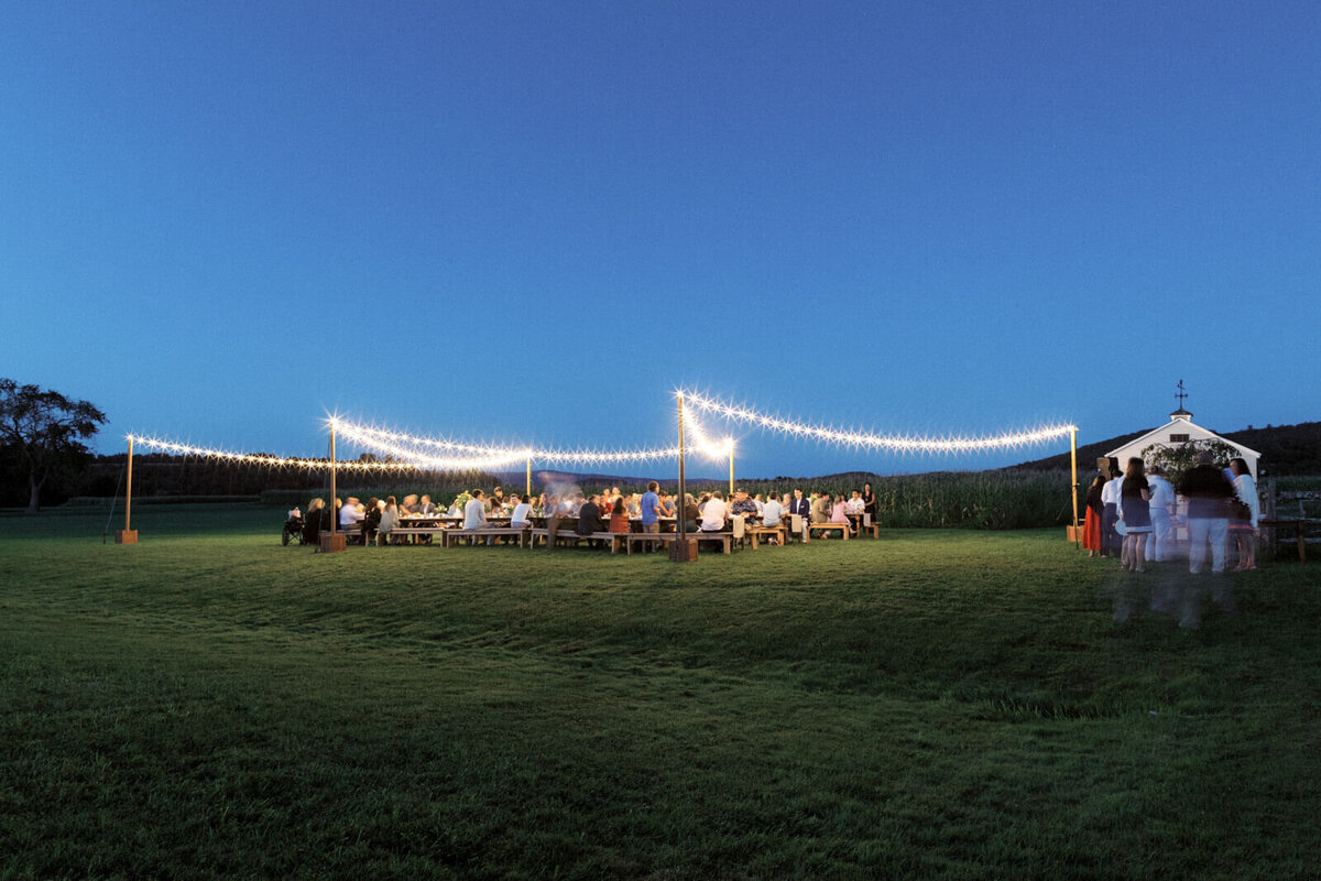 A lot of guests are dining on long tables outdoors, with fairy lights above them, at Lion Rock Farms, CT. Image by Jenny Fu Studio