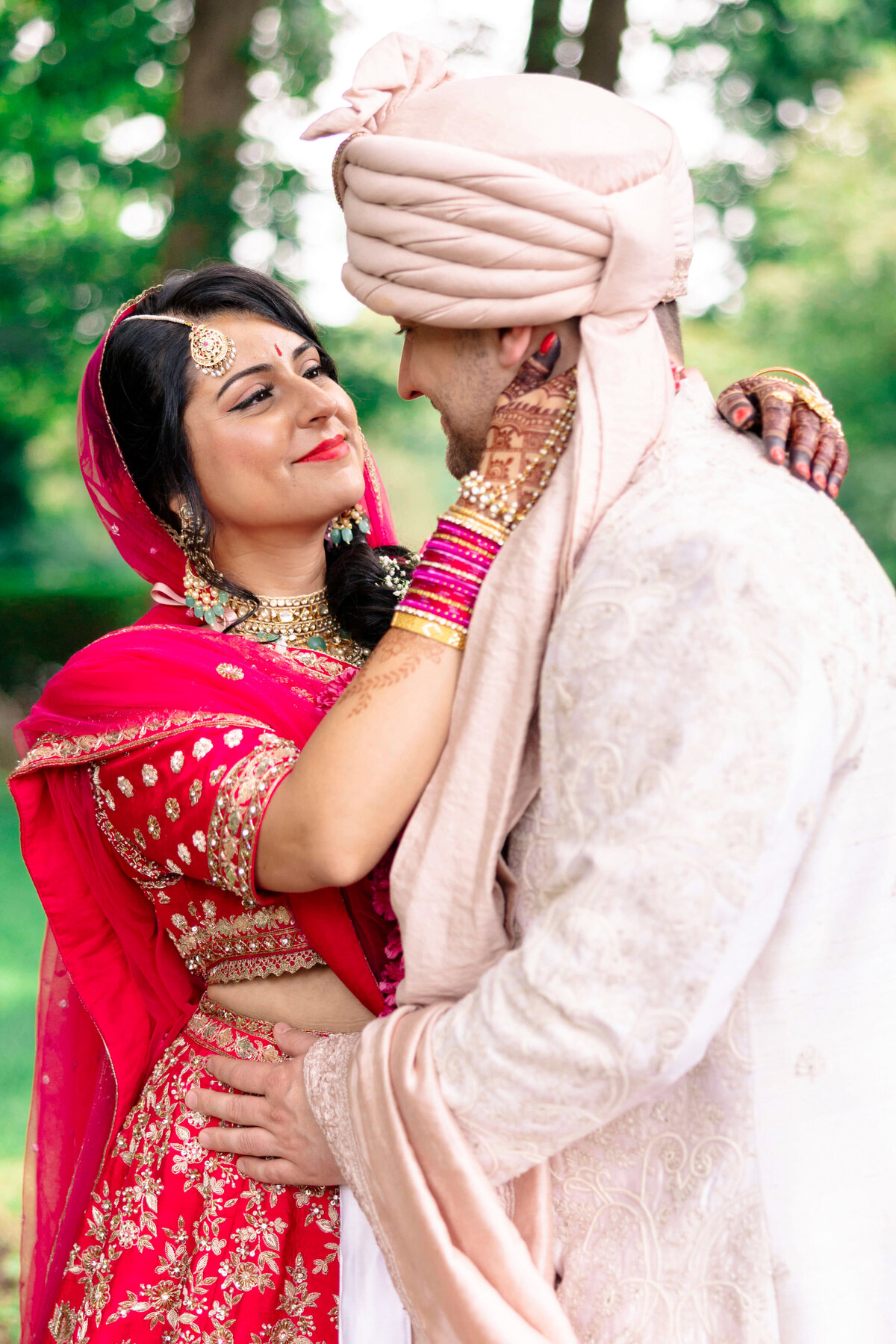 A couple in traditional South Asian wedding attire gazes lovingly at each other. The woman wears a red lehenga with gold embroidery, and the man is in a white sherwani with a beige turban. They are standing outdoors with greenery in the background.