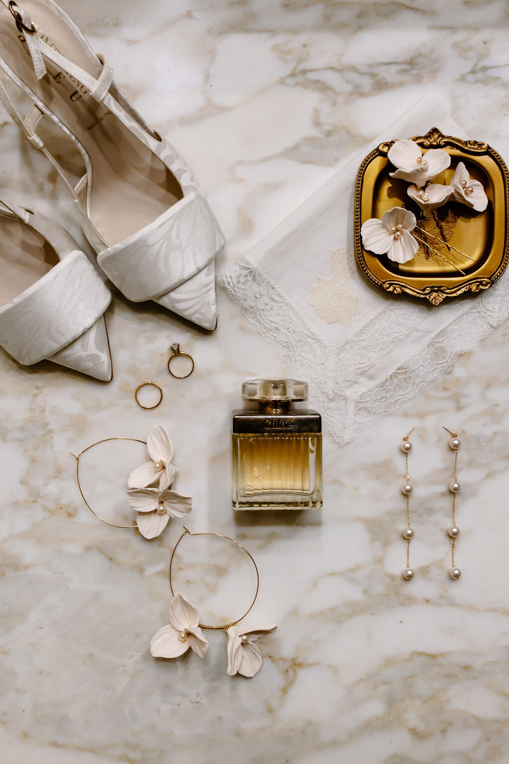 Bird's eye view flatlay photo of elegant bridal shoes, earrings and perfume lay across marble countertop.