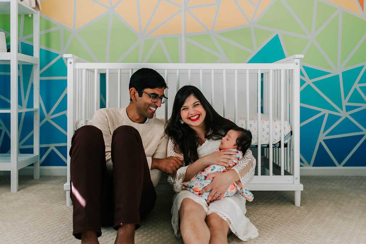 parents holding baby girl in front of crib and laughing during their newborn photos