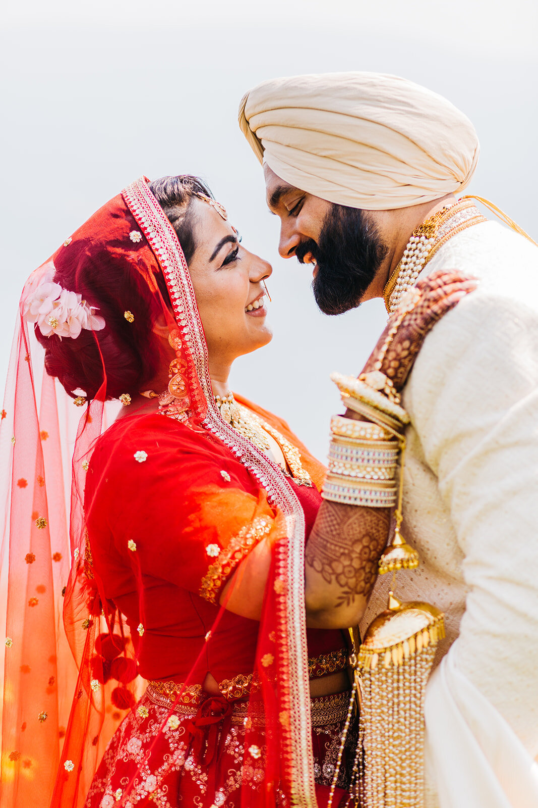 Sikh bride with a red veil and groom in a beige suit look into each other's eyes and smile.